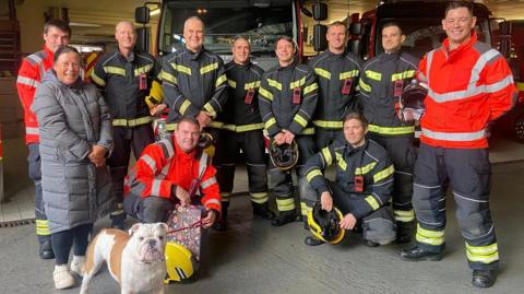 An English Bulldog called George Bullfrog stands next to his owner Sandra, who is wearing a large grey coat, and the Jersey Fire Service crew who rescued him after a cliff path fall. The crew are wearing their overalls and one of the firefighters has a bag which has been given to the crew as a thank you gift by Sandra and George.
