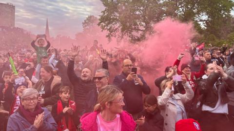 York City fans cheer on their team as an open top bus passes. Red flare smoke fills the air.