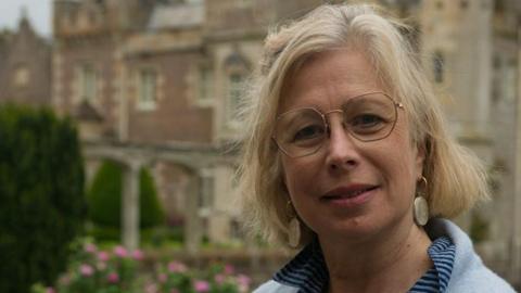A woman with short, blonde hair and wire-rimmed glasses in a light blue jumper with a blue striped blouse underneath stands in front of Abbotsford House the former home of Sir Walter Scott