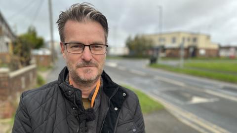 Chris Watts looks at the camera, wearing glasses and a dark coat, in front of one of Swindon's Roads