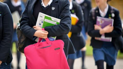 Unidentifiable students carrying bags and books at an unnamed secondary school. The students are wearing navy blue blazers and blue skirts. 