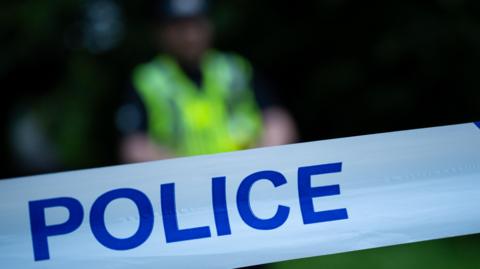 In the foreground is a close up of white police tape with the word POLICE in blue. In the background is a blurry image of a police officer in uniform.