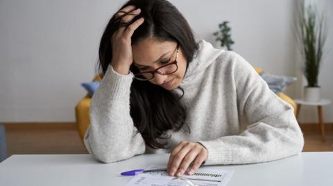 A stock photo of a dark-haired woman with glasses sitting at a table with a bill in front of her. She has a fatigued expression and rests her head on one hand.