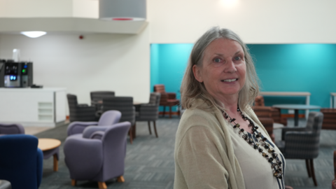 Catherine Lamond looks at the camera, standing side-on in a room with white and blue walls. Chairs and tables are situated behind her. She wears a light top and cardigan and dark beaded necklace.