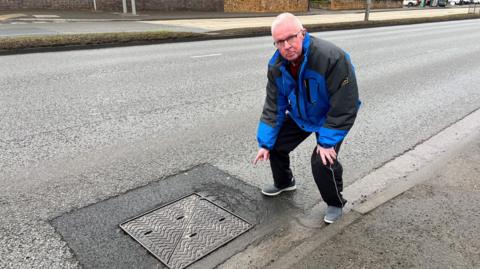 A man pointing at a noisy manhole cover on Derby Road in Beeston 