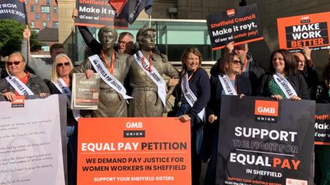 A group on men and women stand next to a statue of two female steelworkers. The group are holding orange and black signs, one of which says 'support the fight for equal pay in Sheffield'.