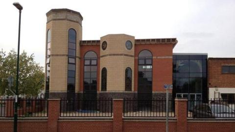 Exterior of the Mosque building with a tall round-shaped side pillar and glass windows - with a parking lot in front and a black grill on top of a red brick wall boundary.