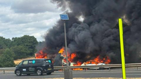 Two flaming vehicles viewed through the central reservation barrier