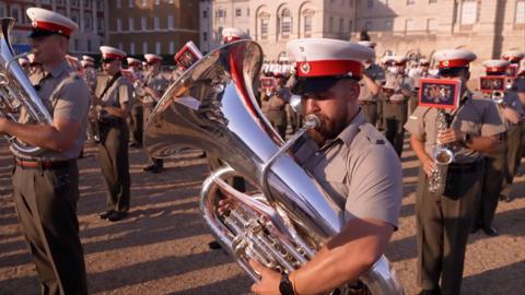 A marching band playing outside a building with a man playing a trombone in foreground