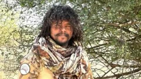 A man in camouflage standing in front of a tree