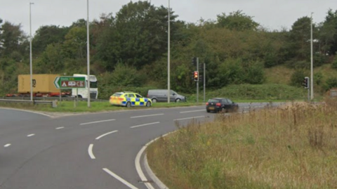 A Google Maps street view image of the Dock Spur roundabout in Felixstowe. A police car is parked up on the left and a black car is parked at the traffic lights. In the distance is a grey van and a large white and yellow lorry.
