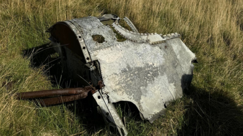 A rusty silver ands bronze wreckage of a cockpit lying on green grass