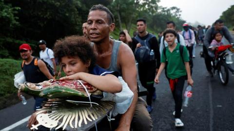 A father and daughter ride a bicycle amongst a group of migrants on the road, making the way to the US Border