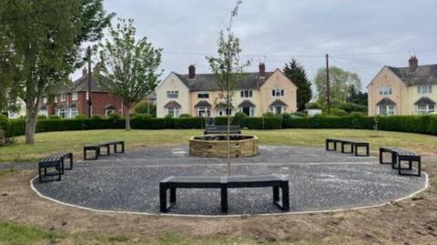 A green enclosed space on an estate. In the middle is a circular bit of tarmac with black benches dotted around it and a circular wooden pit in the centre.