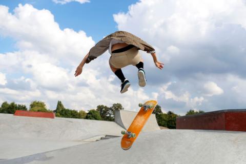A stock image of a man flying through the air at a skatepark with his orange skateboard beneath him.