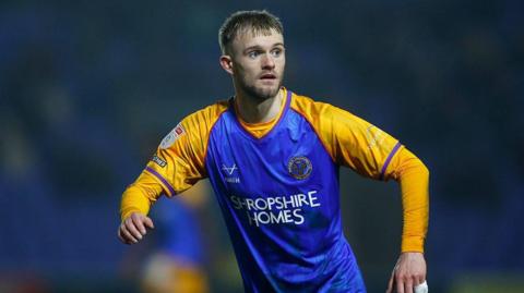 Taylor Perry runs in midfield, looking to his left, wearing Shrewsbury home kit of yellow and blue