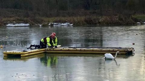 Large lake with two firefighters crouching on an inflatable path to reach a swan stuck in ice