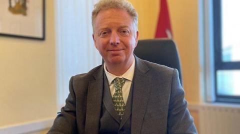 Alex Allinson sitting at his office desk with a standing Isle of Man flag in the background. He is wearing a grey suit and waistcoat with a green patterned tie and white shirt. 