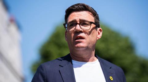 Andy Burnham has dark brown hair, dark brown glasses, a white T-shirt and blue jacket.