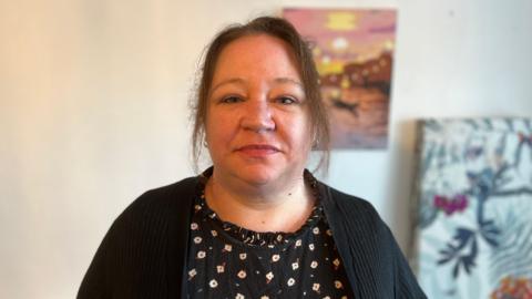 Jo Barnes looks at the camera in the living room of the property she is temporarily renting. She has brown hair and wears a black cardigan with a navy top with white flowers on underneath