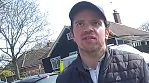 Bodycam image of Luke D'Wit being arrested. He's wearing a hat and padded jacket