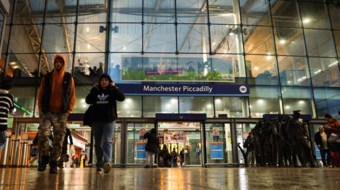 People walk out of the glass-fronted entrance to Manchester Piccadilly train station on a wet day.