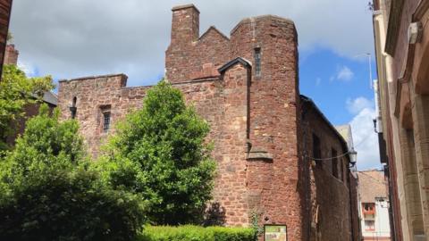 An ancient red brick monastery type building, with trees and bushes in the foreground