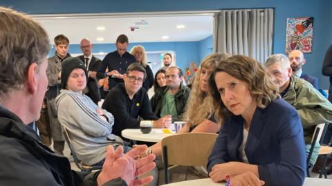 Wayne, a user of Salford charity Loaves and Fishes sat a table in a black jacket with a grey hood, speaking to a room of people. Work and pensions secretary Liz Kendall sits with her arms crossed across from Wayne, listening on the same table.