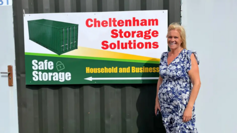 A woman with blonde hair and a blue and white floral dress smiles as she stands in front of a mostly green and white sign with red text 