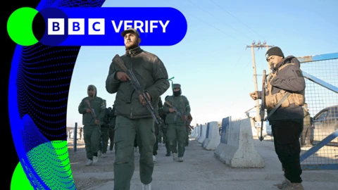 Men armed with AK-47-style rifles and wearing green military clothing walking inside a camp