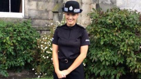 A woman with blonde hair smiling. She is wearing a police uniform and standing in front of green bushes.