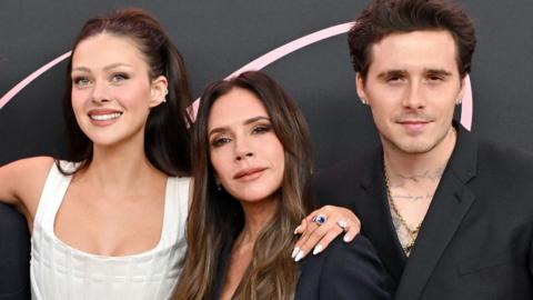  Nicola Peltz Beckham, Victoria Beckham and Brooklyn Peltz Beckham stand together at a press event . Victoria Beckham is in the middle. Nicola has her arm around Victoria. They are in evening dress
