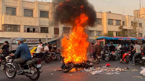 People gathering next to a burning motorcycle during protests in Tehran on the 8th October
