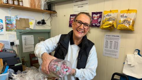 A woman wearing glasses smiles as she pours sweets out of a plastic container.