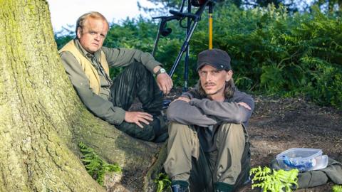 Two men sit at the base of a large tree. They are wearing long trousers and shirts. One has a blue cap on. 