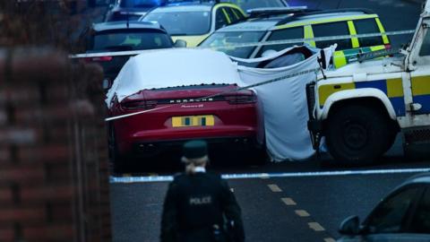 A red Porsche car is covered in a white forensic sheet and surrounded by a white, blue and yellow PSNI Landrover, a green and yellow ambulance vehicle and another emergency response vehicle. A police woman is in the foreground of the image.