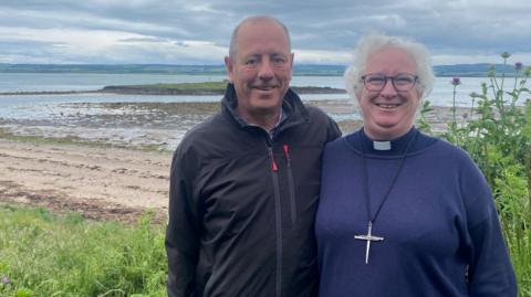 Dr Richard Hills and his wife Dr Sarah Hills are a couple in their sixties. She is wearing a purple top with a clerical collar and a large cross necklace. She has curly grey hair and glasses. He is wearing a zipped-up dark outdoor jacket and has short receding grey hair. Both are smiling. Behind them is a beach with a small low island in the sea and, in the distance, a larger stretch of rolling land across the water.
