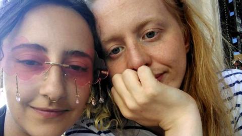 A selfie of couple Daisy, 24 and Ramona, 25. Daisy has her dark hair pulled up, and is wearing pink tinted sunglasses with jewels dangling underneath them and has a nose ring piercing. Ramona has long, straight blonde hair, and is wearing a white T-shirt with horizontal navy stripes. She also has her hand resting on her chin blocking her mouth.