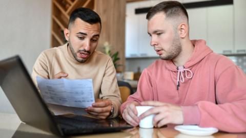A young couple, two men, look at an electricity bill and a laptop in the kitchen of their home