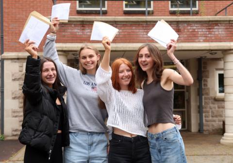 Four girls standing outside a building, holding their A-Level results above their heads