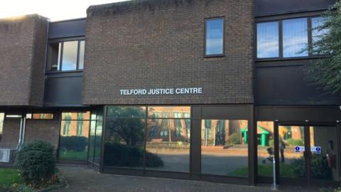 The front of a dark brown brick building with large glass windows. The words Telford Justice Centre are on the wall above the windows.