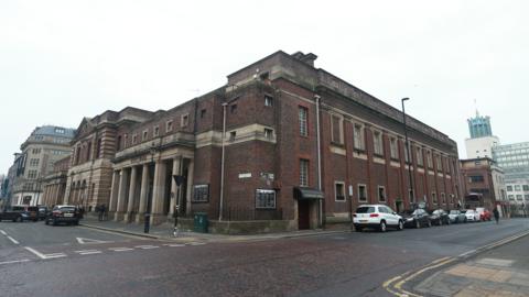 A picture of Newcastle City Hall. The photograph is taken from the corner of the street so two sides of the red-brick building are in view. The entrance stands to the left and has large sandstone pillars in front of the door. The City baths are further to the left.