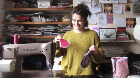 Emma Crick holding a reusable mentrual pad in each hand while standing behind a sewing machine