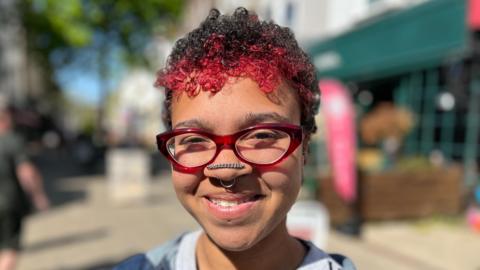 A woman with short, black and red curly hair. She is wearing red-frame glasses and has a septum piercing and a chain across the front of her nose.
