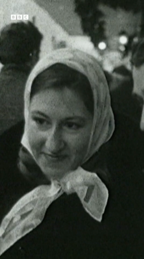 A black and white still from a TV news programme of a woman standing in a shopping street with a few people behind her. It is a head and shoulders shot and she is wearing a silk scarf over her head and smiling.