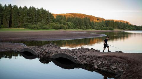 View of a depleted reservoir with a boy walking across a bridge with a fishing rod
