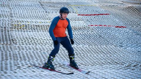 An older gentleman skiing down an artificial snow slope. He is wearing a helmet, a blue and orange top and black gloves.