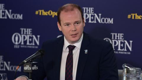 Lyons is sitting in front of a mic talking to the media. The backdrop behind him is for the Open. He's wearing a navy suit with a white shirt and a tie. A jug of water is sitting on the table next to him.