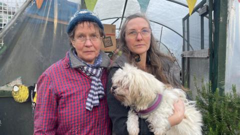 Two women stand together in front of a plastic green house. One of the women is holding a white dog