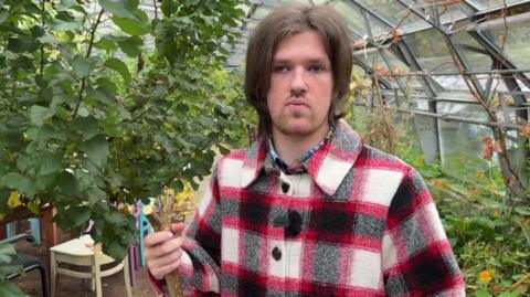 Adam has mid length brown hair and is wearing a red and black checked jacket and is stood in a greenhouse surrounded by plants